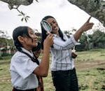 Students in school uniforms explore nature with a teacher using a magnifying glass.