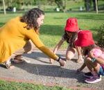 A nanny and two children drawing with chalk on a sunny day outdoors.